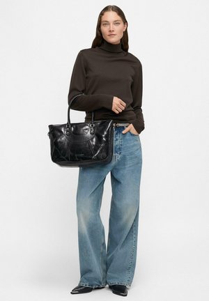 Donna che indossa un dolcevita scuro e jeans a gamba larga, che tiene in mano una grande borsa nera in pelle con doppio manico.