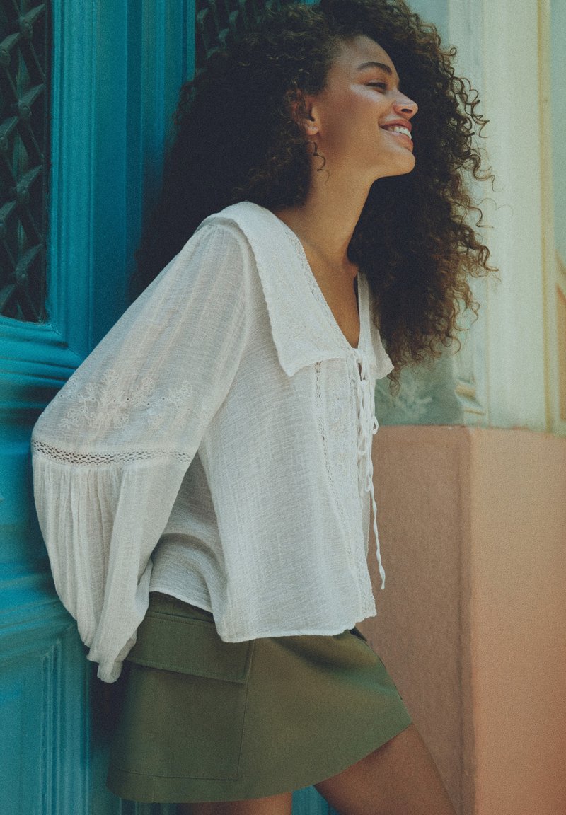 Femme aux cheveux bouclés souriante, portant un chemisier blanc avec des détails en dentelle et une jupe vert olive, appuyée contre une porte bleue.