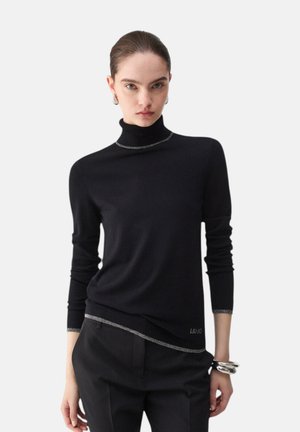 Donna con i capelli lisci tirati indietro che indossa un maglione nero a collo alto con rifiniture argentate e pantaloni neri, accessoriata con braccialetti e orecchini argentati.