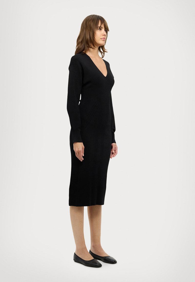 Rochie tricotată, neagră, cu mânecă lungă, cu decolteu în V, siluetă ajustată, textură canelată, lungime până la genunchi și asortată cu pantofi negri cu talpă plată.