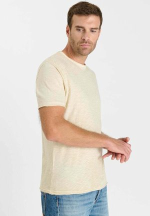 Hombre vistiendo una camiseta beige de manga corta y vaqueros azules, mirando hacia abajo con las manos entrelazadas frente a un fondo blanco liso.