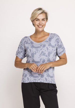 Smiling woman with short blonde hair wearing a gray patterned short-sleeve shirt and black pants, standing with hands clasped.