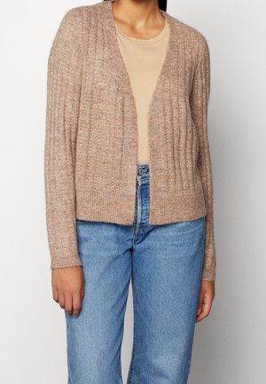 Nainen, joka on pukeutunut beigeksi neulotun ribbikardiganin päälle vaalean ruskeaan t-paitaan ja sinisiin korkeavyötäröisiin farkkuihin, seisoo yhtenäistä taustaa vasten.