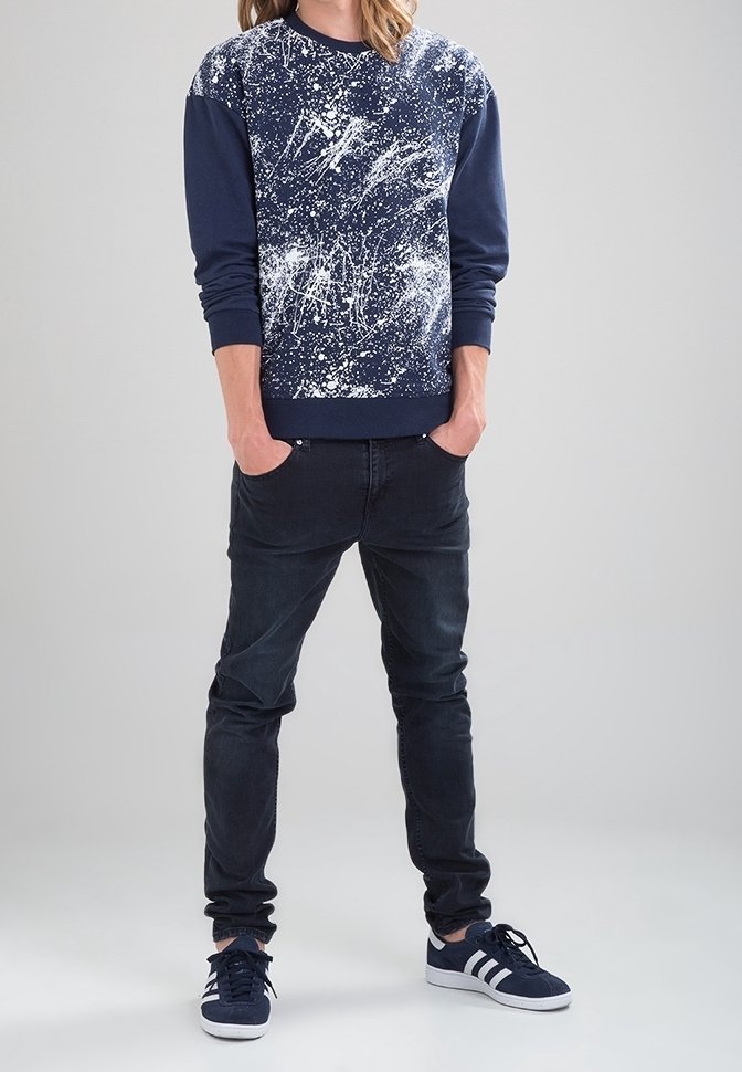 Sudadera azul marino con diseño de manchas blancas, combinada con jeans oscuros y zapatillas azul marino con rayas blancas. Ajuste casual y estilo moderno.