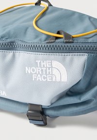 Τμήμα τσάντας North Face ανοικτού γαλάζιου χρώματος με φερμουάρ, λευκό κεντημένο λογότυπο, μαύρη αγκράφα και κίτρινη θηλιά κορδονιού στην κορυφή.