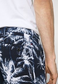 Homme portant un short bleu marine à imprimé palmiers blancs et une chemise blanche, montrant les détails de la poche latérale et arrière.
