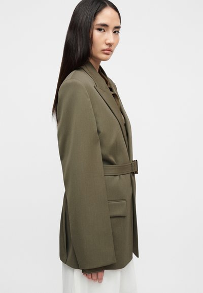 Λαδί πράσινο σακάκι με ραφινάτο κόψιμο, ζώνη στη μέση, κοφτερά πέτα και λεπτομέρεια μπροστινής τσέπης. Ομαλή υφή με δομημένη εφαρμογή.