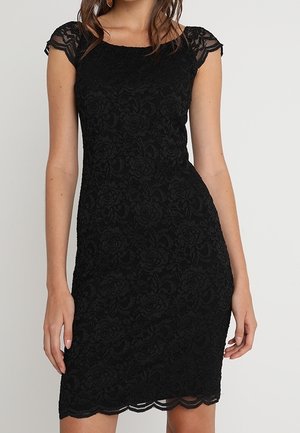 Schwarzes knieumspielendes, figurbetontes Spitzenkleid mit Kappenärmeln und gezacktem Saum, getragen von einer Frau, die vor einem einfarbigen Hintergrund steht.