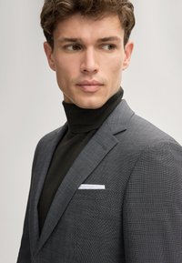 Grau blazer aus strukturiertem Stoff, maßgeschneiderte Form und mit Reverskragen. Getragen über einem dunklen Rollkragenpullover, mit einem weißen Taschentuch.