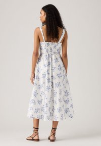 Donna con capelli ricci indossa un abito midi bianco con stampa floreale blu e spalline sottili, abbinato a sandali neri allacciati, voltata di spalle.