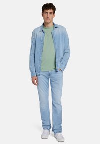 Camicia in denim azzurro chiaro indossata sopra una maglietta verde, abbinata a jeans azzurro chiaro e sneakers bianche; stile casual, vestibilità rilassata e chiusure a bottoni.