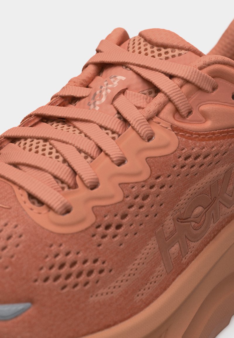 Gros plan d'une basket HOKA de couleur corail avec un mesh texturé, des lacets épais et un design ondulé des œillets sur le panneau latéral.