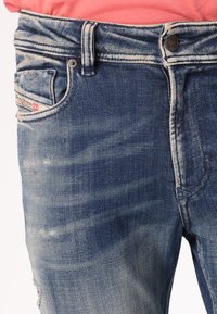 Traperice u srednje plavoj boji s izbljeđenim dijelovima i istrošenim detaljima. Imaju brendiranu stražnju džepu i tradicionalni dizajn s pet džepova.