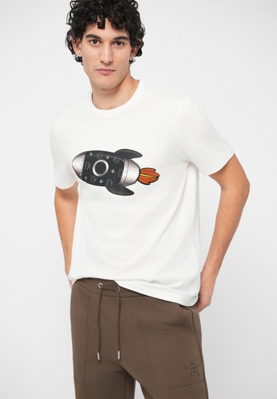 Νεαρός άνδρας με σγουρά μαλλιά που φοράει λευκό μπλουζάκι με μαύρη γραφική απεικόνιση πυραύλου και καφέ παντελόνια με κορδόνι στη μέση, με τα χέρια στις τσέπες.