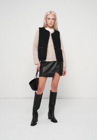 Černá beránková vesta přes béžový svetr, doplněná černou koženou sukní a černými kozačkami po kolena. Modelka drží malou černou kabelku.