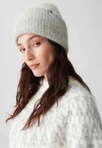 Light gray knit beanie made from soft, textured yarn, featuring a small logo tag. Worn with a patterned white sweater.