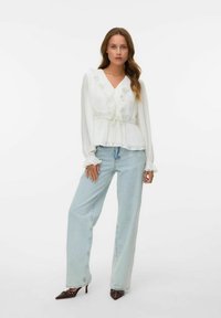 Hvit, plisserte bluse med volangkant ved halsen og mansjettene; parret med lyseblå vide jeans og spisse brune hæler.