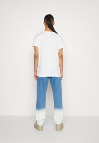 Osoba s dlouhými vlasy v culíku stojící zády, oblečená v bílém tričku, modro-bíle přechodových džínách a béžových botách.