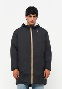 Veste longue noire avec capuche, dotée d'un zip jaune et blanc et d'un logo sur la poitrine. Intérieur en polaire pour plus de chaleur.