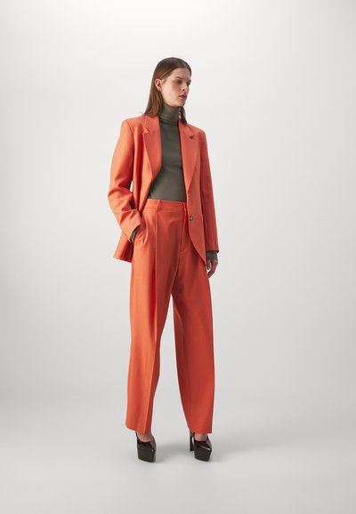 Sacou portocalie croit cu pantaloni largi, asortați cu un pulover verde măslin. Țesătură netedă cu un design structurat și detalii cu nasturi aurii.