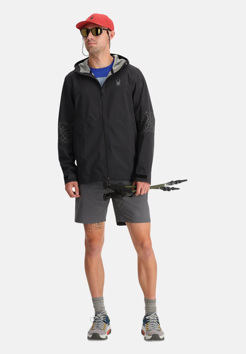 Czarna kurtka z szarym wykończeniem i rękawami w siatkowy wzór, w zestawieniu z szarymi szortami. Na głowie czerwona czapka i okulary przeciwsłoneczne, trzyma trekkingowe kijki.