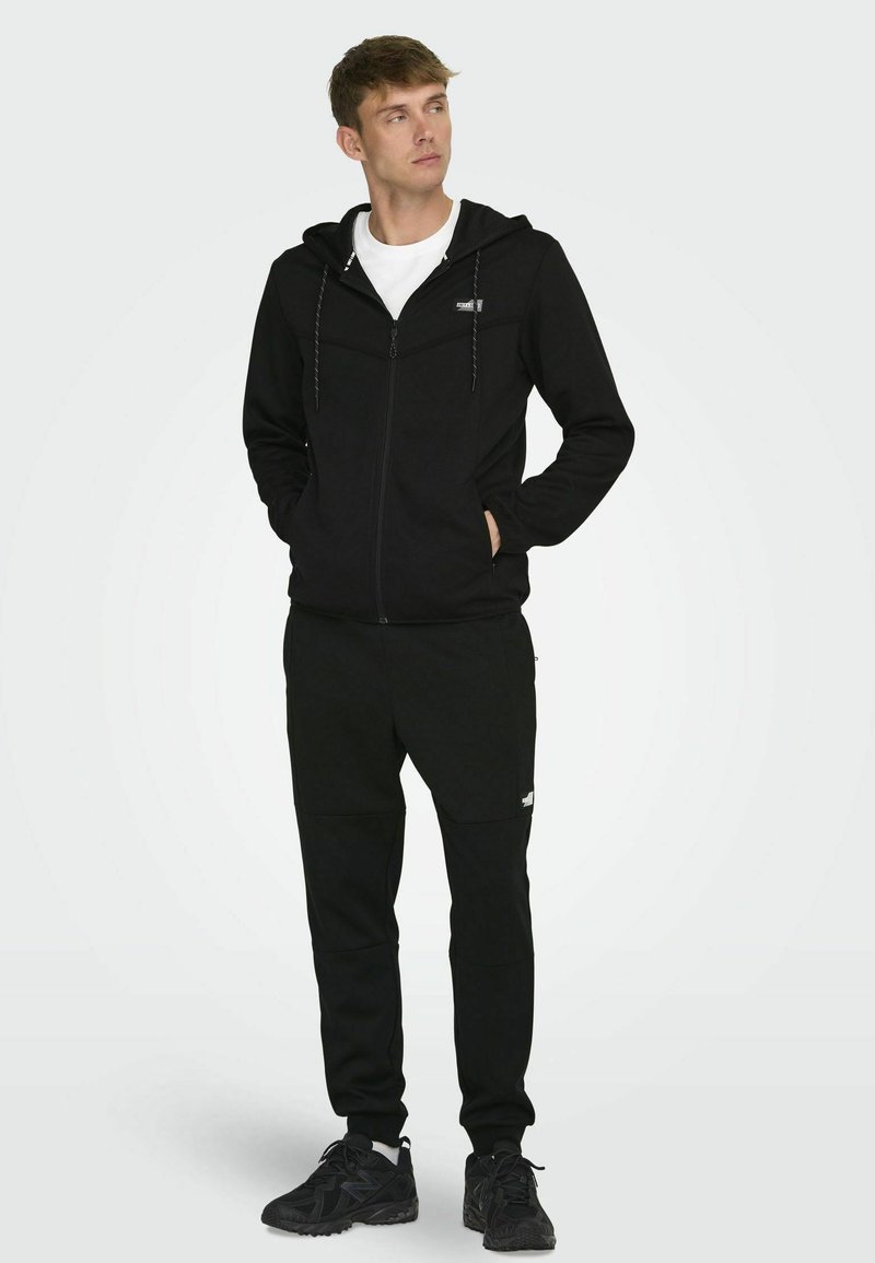 Zwarte zip-up hoodie met een trekkoordcapuchon en wit logo, gecombineerd met bijpassende zwarte joggers gemaakt van zachte stof en met textuurdetails.