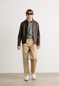 Brun läderjacka, grön tröja, beige byxor, vita sneakers med ljusbruna sulor; ämnet bär solglasögon och står mot en neutral bakgrund.