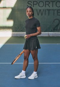 Camiseta atlética negra y falda, sosteniendo una pelota de tenis verde y una raqueta naranja. Zapatillas blancas con suelas texturizadas, en una pista azul.