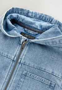Veste en denim bleu clair, dotée d'une fermeture éclair, d'une capuche réglable, d'une poche poitrine et de détails de surpiqûres visibles. Étiquette de marque visible.