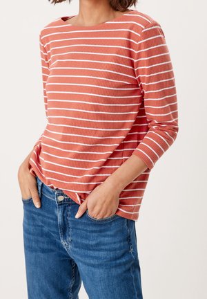 Žena v oranžovo-rustikální a bíle pruhované košili s dlouhým rukávem lehce zastrčené do modrých džín s rukama v kapsách na jednobarevném pozadí.