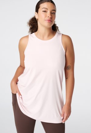 Vrouw met krullend haar in een lichtroze mouwloos topje en donkere legging, staand met één hand in haar zak tegen een effen achtergrond.