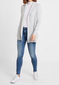 Szary dziergany kardigan z otwartym przodem, długimi rękawami, noszony na jasnym golfie i niebieskich obcisłych dżinsach, zestawiony z białymi trampkami.