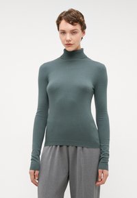 Grønn genser med høy hals i en tettsittende design, rillede detaljer ved halsen og lange ermer; kombinert med grå, plisserte bukser. Glatt tekstur.