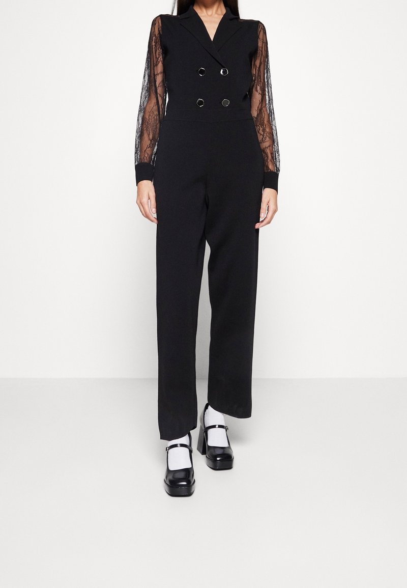 Femme portant une combinaison noire croisée à manches longues en dentelle transparente et des chaussures Mary Jane à plateforme noires avec des chaussettes blanches.