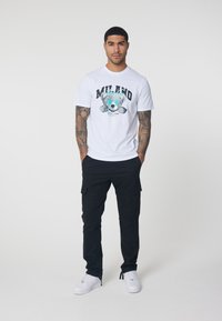 White cotton t-shirt with a graphic featuring a koala and "MILANO" text. Paired with black cargo pants and white sneakers.