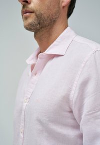 Camisa de lino de un suave tono rosa, con un cuello relajado, botones en la parte frontal y un sutil logotipo bordado en el pecho. Tejido ligeramente texturizado.