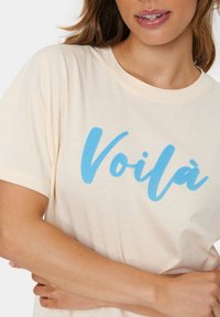 Šviesiai kreminė marškinėliai, pagaminti iš minkštos medžiagos, su žodžiu "Voilà" ryškiu mėlynu kursyvu per visą priekinę dalį. Matomos trumpomis rankovėmis.