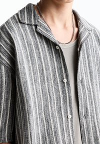 Chemise à manches courtes rayée gris et blanc avec col, dotée d'une fermeture à boutons et d'un tissu texturé, portée sur un débardeur beige.