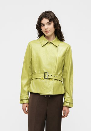 Frau mit welligem dunklem Haar, die eine hellgrüne texturierte Jacke mit Gürtel und dunkelbraune Hose trägt, steht vor einem schlichten hellen Hintergrund.