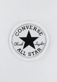 Vitt tygmärke med en svart stjärna, med texten "CONVERSE ALL STAR" och "Chuck Taylor" i svart broderi samt en sydd kant.