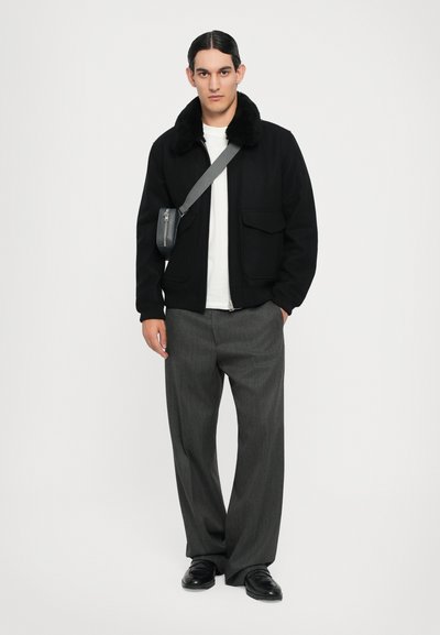 Schwarze Jacke mit einem Pelzkragen, weißes T-Shirt, graue Plisseehose und schwarze Schuhe. Kleine graue Umhängetasche, die über der Schulter getragen wird.