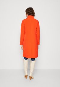 Orange tætsiddende frakke med høj krave, lange ærmer og en lige snit, kombineret med knæhøje cremefarvede støvler og denimjeans.