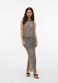 Donna che indossa un top trasparente senza maniche a motivo zigzag e una gonna maxi coordinata con spacco laterale, abbinati a sandali marroni su sfondo bianco.