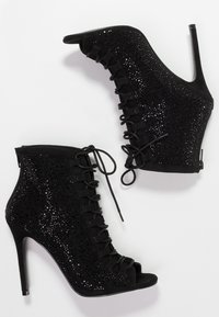 Zwarte enkellaarsjes met hoge hak en open teen, voorzien van gestructureerde stof, veters aan de voorkant en glinsterende versieringen aan de zijkanten.