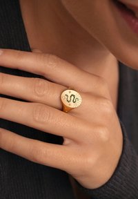 El anillo de oro presenta un diseño circular con una serpiente enrollada en el centro, rodeada de motivos de estrellas y líneas radiantes, con una textura suave.