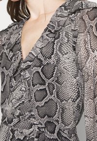 Schwarze und weiße Schlangenlederdruck-Bluse mit rüschigem Ausschnitt und langen Ärmeln, hergestellt aus einem transparenten, leichten Stoff mit texturiertem Muster.