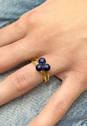 Main avec une bague double anneau en or ornée de trois perles rondes bleu foncé, reposant sur un tissu en denim bleu clair.