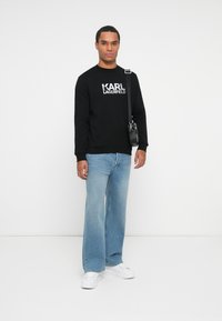 Sudadera negra con el logo blanco "KARL LAGERFELD", combinada con unos vaqueros de pierna ancha azul claro y zapatillas blancas. Incluida una mochila cruzada negra.