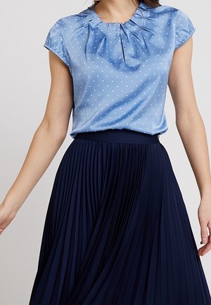 Vrouw in een lichtblauwe, korte mouwen, gestippelde blouse gestoken in een hoge taille, marineblauwe plissé rok.