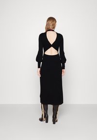 Schwarzes, langärmeliges Kleid mit taillierter Silhouette, ausgestattet mit einem Cutout im Rücken und strukturiertem Stoff. Kombiniert mit knielangen schwarzen Stiefeln.